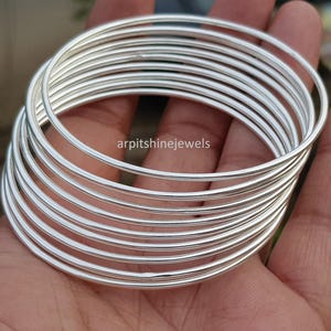 Puede incluir: Una pila de pulseras plateadas, cada una con un diseño liso y redondeado. Las pulseras están apiladas unas sobre otras, creando un efecto de capas. Las joyas se muestran en una mano. El texto "arpitshinejewels" es visible.