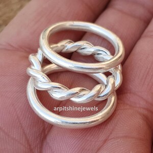 May include: Three silver rings interlocked, featuring a braided design on the connecting bands. The rings are smooth and shiny, reflecting light. The text "arpitshinejewels" is visible on the bottom ring.