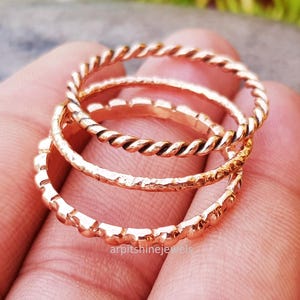 May include: A close-up shot of three rose gold rings. The top ring features a twisted rope design, the middle ring has a hammered texture, and the bottom ring has a beaded edge. The rings are stacked together.