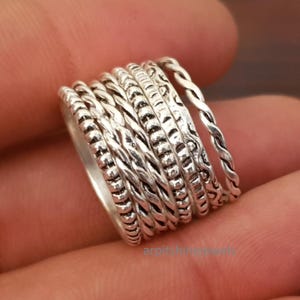 May include: A stack of silver rings featuring diverse textures such as braided, beaded, and patterned designs. The rings are displayed together, highlighting the unique styles and intricate details of each piece. The rings are made from a shiny, metallic material.
