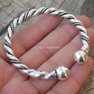 Puede incluir: Una pulsera de plata con un diseño de cuerda trenzada. La pulsera tiene un extremo sólido y redondeado en cada lado. El texto "arpitshinejewels" es visible.