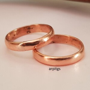 Pode incluir: Dois anéis cor de cobre são exibidos, um sobrepondo-se ligeiramente ao outro. Os anéis têm um design liso e arredondado. Os anéis estão sobre uma superfície de cor clara. O texto "arpitgs" é visível.