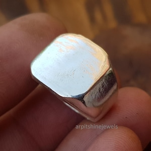 May include: A close-up of a silver ring with a square face and rounded edges. The ring is highly polished and reflects light. The text "arpitshinejewels" is visible at the bottom of the image.