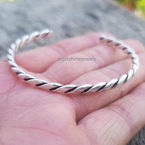 Op de afbeelding: Een zilverkleurige manchetarmband met een gedraaid touwontwerp. De open manchetarmband wordt op een hand getoond. De armband heeft een getextureerde, gevlochten uitstraling. De sieraden zijn een eenvoudig, elegant accessoire.