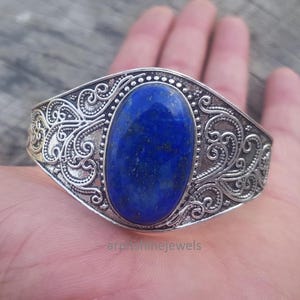May include: Silver-toned cuff bracelet featuring an oval, deep blue gemstone. The bracelet has an ornate, swirling filigree design around the gemstone and along the band. The jewelry is displayed on a hand.