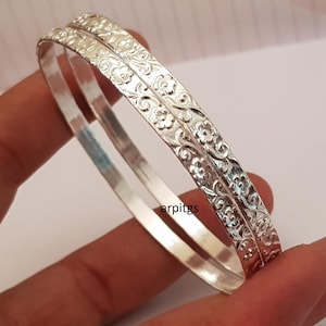 May include: Two silver bangles with floral patterns. The bangles have a smooth inner surface and an embossed design of flowers and leaves on the outer surface. The bangles are held in a hand.