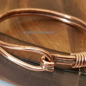 May include: A copper-colored bangle bracelet with a unique clasp design. The bracelet is made of a single piece of metal that is bent and shaped into a circular form. The clasp is a hook and eye design. The bracelet is displayed on a wooden surface.