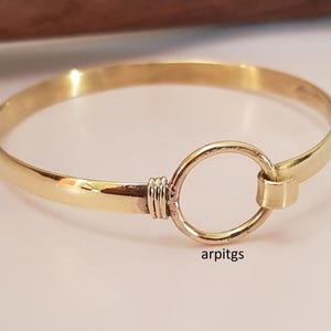 May include: A gold-colored bangle bracelet with a circular clasp. The bracelet has a smooth, polished finish with a connecting ring clasp. The word "arpitgs" is printed on the bracelet.
