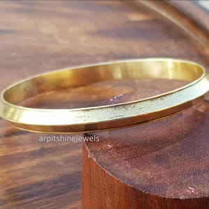 Puede incluir: Una pulsera de brazalete dorada con un diseño plano y angular. La pulsera está sobre una superficie de madera. El texto "arpitshinejewels" es visible en la superficie. Esta joya es adecuada para el uso diario.