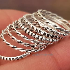 May include: A selection of silver rings with diverse textures. Some rings have a twisted rope design, while others feature small, dark rectangular patterns. The rings are stacked, displaying their different styles. Each ring is a unique design.
