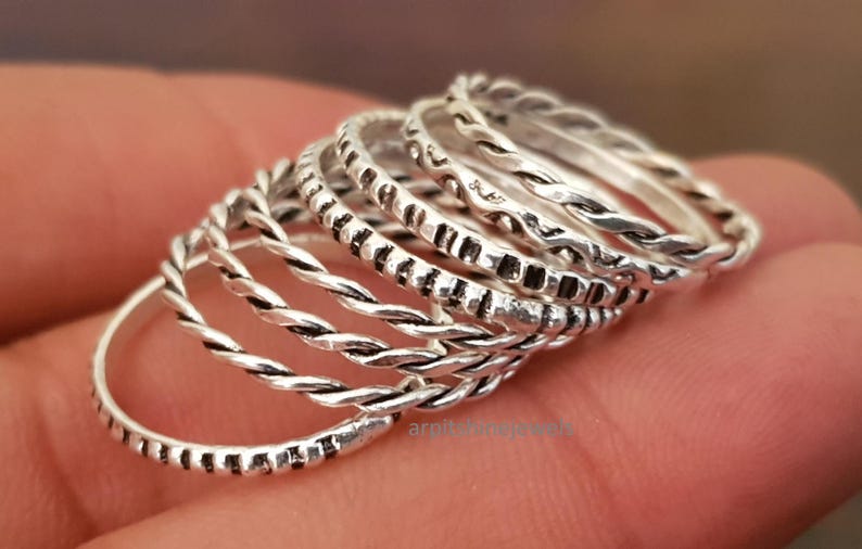 May include: A selection of silver rings with diverse textures. Some rings have a twisted rope design, while others feature small, dark rectangular patterns. The rings are stacked, displaying their different styles. Each ring is a unique design.