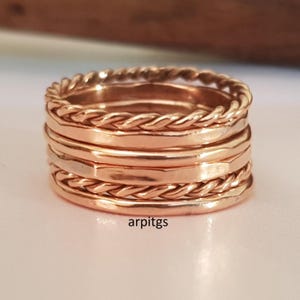 Op de afbeelding: Een stapel ros&eacute;gouden ringen. De ringen hebben een mix van texturen, waaronder een gevlochten ontwerp en gladde banden. De ringen zijn op elkaar gestapeld, waardoor een gelaagde look ontstaat. De ringen worden gepresenteerd op een wit oppervlak.