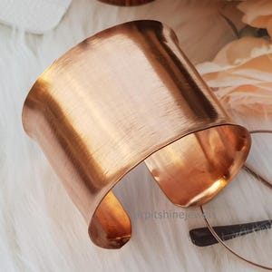 Può includere: Un ampio bracciale a polsino in rame con finitura in metallo spazzolato. Il design aperto presenta una forma curva che si avvolge attorno al polso. Il bracciale è esposto su una superficie bianca e soffice.