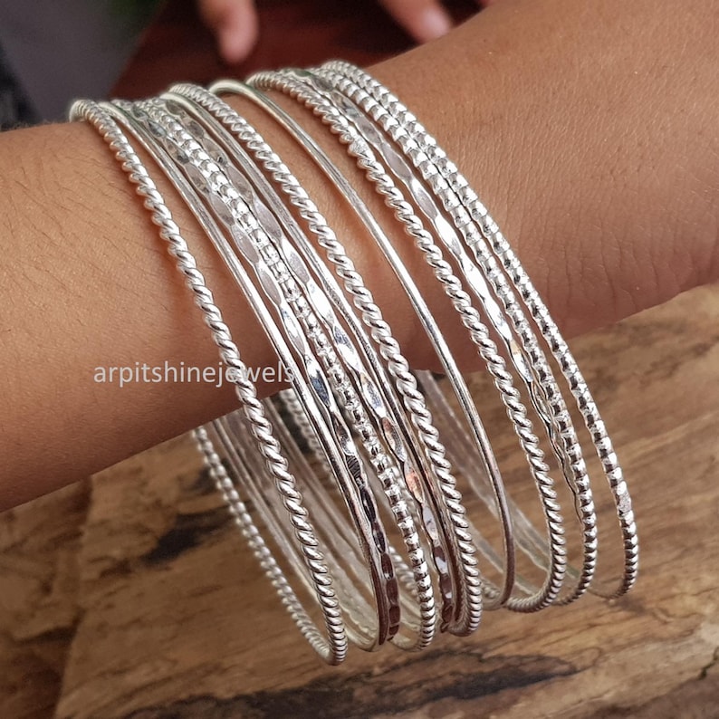 May include: A set of silver-toned bangles featuring a variety of textures, including smooth, twisted rope, and hammered finishes. The bracelets are stacked together, and the text arpitshinejewels is visible on the left side of the image.