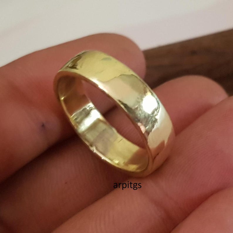 May include: A gold-colored ring with a hammered texture. The ring is a simple band design, reflecting light. The ring is held in a hand. The text "arpitgs" is visible on the ring.