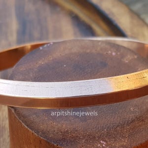 Puede incluir: Una pulsera brazalete de color cobre con un diseño plano y ligeramente biselado. La pulsera se exhibe sobre una superficie de madera, mostrando su brillo metálico. El texto "arpitshinejewels" es visible.