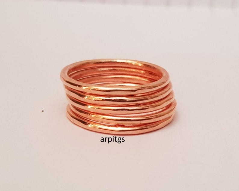 May include: A stack of copper-colored rings. The rings have a hammered texture and are stacked together. The rings are on a white surface. The text "arpitgs" is visible on the bottom right.