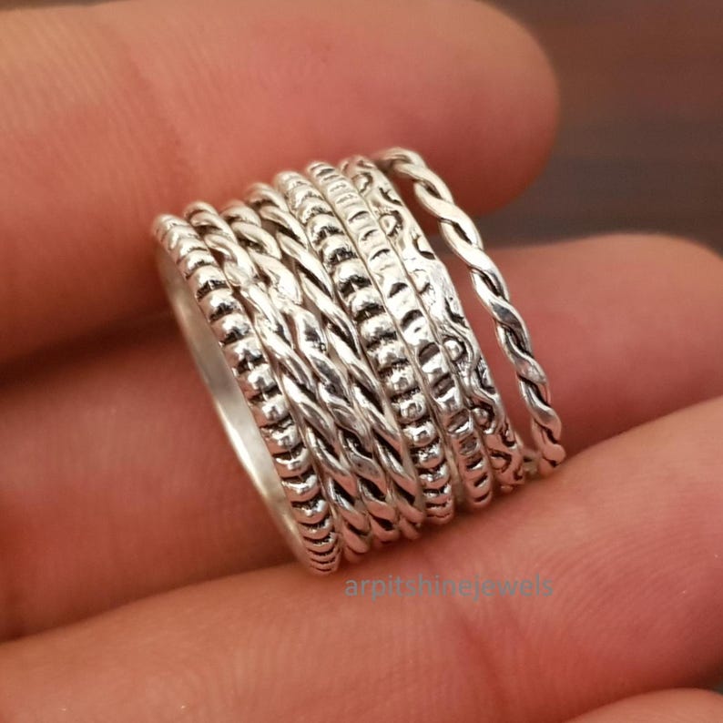 May include: A close-up of a silver ring featuring multiple bands with diverse textures, such as braided, beaded, and patterned elements. The ring's design is intricate, with each band showcasing unique detailing. The ring is likely crafted from sterling silver.