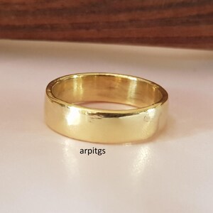 May include: A gold-colored ring with a flat band. The ring has a smooth, polished surface and a simple, classic design. The ring is shown on a light pink surface. The text "arpitgs" is visible on the surface.