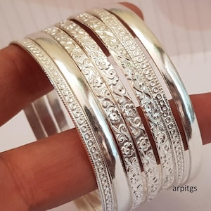 Pode incluir: Um conjunto de pulseiras de prata, algumas com desenhos florais e estampados. As pulseiras estão empilhadas, mostrando suas superfícies polidas e reflexivas. A imagem destaca os detalhes intrincados e o brilho geral das joias.