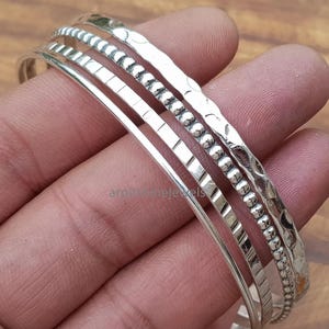 May include: A set of four silver bangles with different textures. One bangle has a hammered finish, another has a beaded design, and the others have linear and smooth surfaces. The bangles are displayed on a hand.
