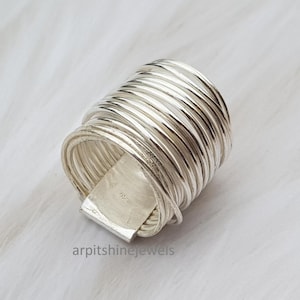 May include: A silver ring with a unique design. The ring is made of multiple layers of thin, coiled metal, creating a textured, wrapped appearance. A rectangular piece of metal is attached to the ring. The ring is displayed on a white, textured surface.