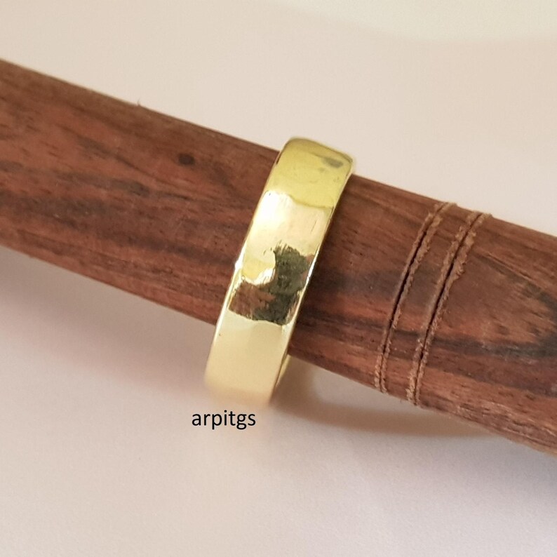 May include: A gold-colored ring with a hammered texture is displayed on a dark brown wooden dowel. The ring has a flat, wide band. The text "arpitgs" is visible at the bottom of the image.