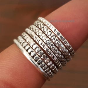 May include: A stack of silver rings with various textured designs, including dotted, ridged, and patterned surfaces. The rings are worn on a finger, showcasing the different styles and the overall aesthetic of the jewelry.