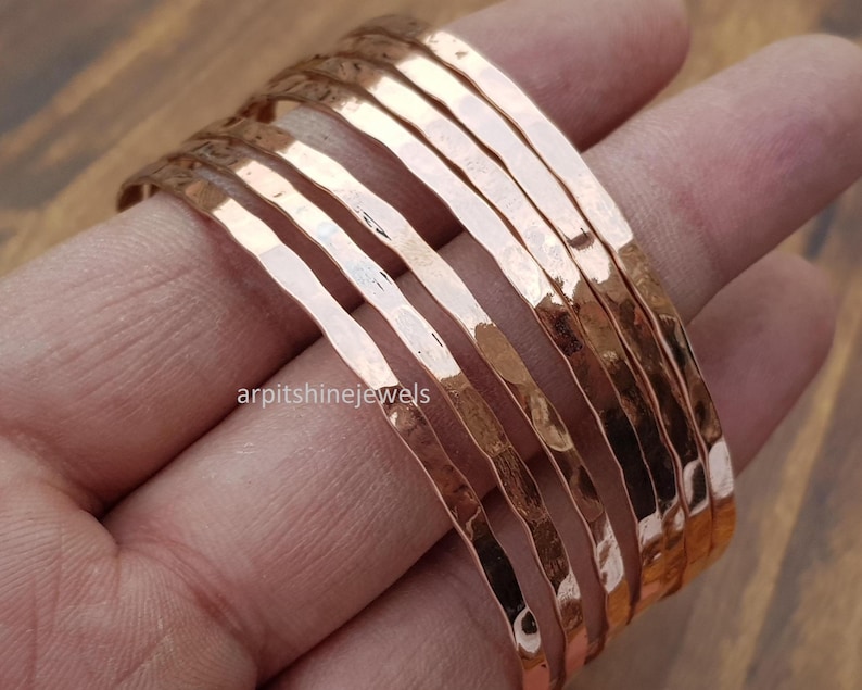 May include: A stack of hammered copper bracelets. The bracelets have a textured surface and a warm, reddish-brown color. The bracelets are stacked on a person's fingers. The text "arpitshinejewels" is visible.