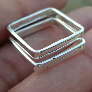 May include: A close-up of a silver ring with a square design. The ring is made of a shiny, reflective metal and has a stacked appearance, with two square bands. The ring is on a person's skin.