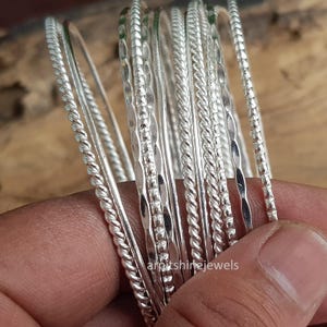 May include: A selection of silver bangles with different textures, including twisted and patterned designs. The bangles are shown against a warm, wooden backdrop. The image highlights the jewellery's shine and detail, ideal for accessorising.