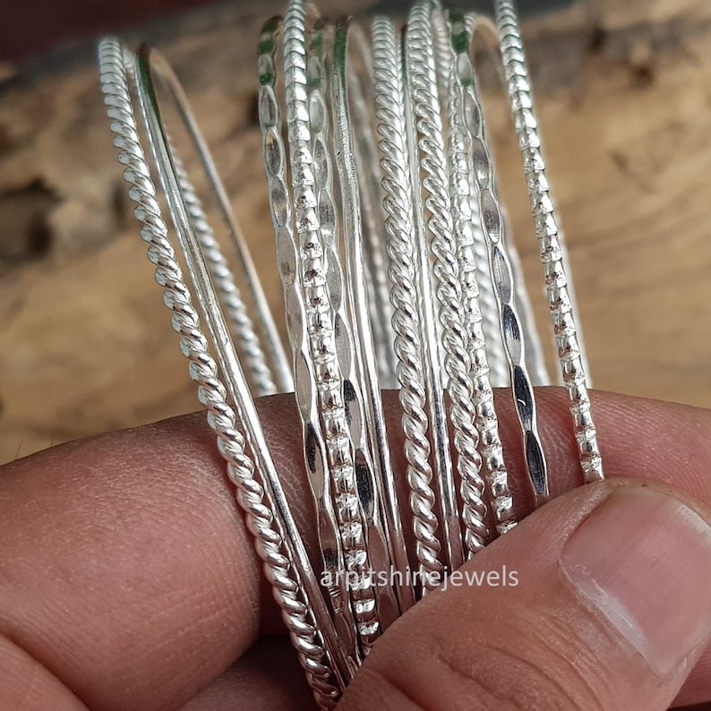 May include: A selection of silver bangles with different textures, including twisted and patterned designs. The bangles are shown against a warm, wooden backdrop. The image highlights the jewellery's shine and detail, ideal for accessorising.