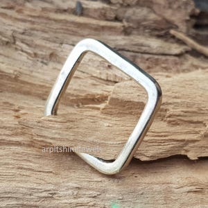 Könnte beinhalten: Ein quadratischer Silberring, der auf einer strukturierten Holzoberfläche liegt. Der Ring hat ein schlichtes, geometrisches Design mit poliertem Finish. Dieses minimalistische Schmuckstück ist ein stilvolles Accessoire.
