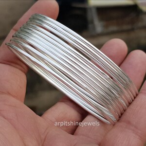 May include: A stack of silver-colored bangles held in a hand. The bangles are thin and round, with a shiny, reflective surface. The jewelry is displayed against a blurred background.