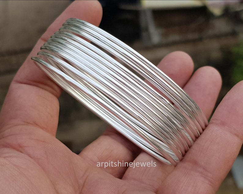 May include: A stack of silver-colored bangles held in a hand. The bangles are thin and round, with a shiny, reflective surface. The jewelry is displayed against a blurred background.