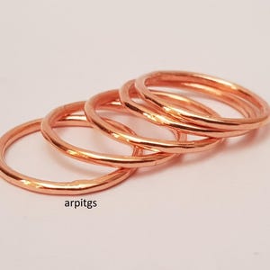 May include: Four copper-colored rings are stacked together, each with a hammered texture. The rings are displayed on a light pink surface. The rings are a simple, minimalist design.