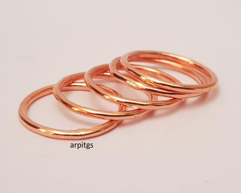 May include: Four copper-colored rings are stacked together, each with a hammered texture. The rings are displayed on a light pink surface. The rings are a simple, minimalist design.