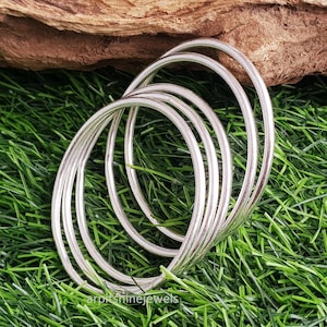 Puede incluir: Un conjunto de pulseras plateadas, dispuestas en una pila suelta, sobre un lecho de hierba verde. Las pulseras son lisas y reflectantes, con un diseño sencillo y elegante. Un trozo de madera está en el fondo.