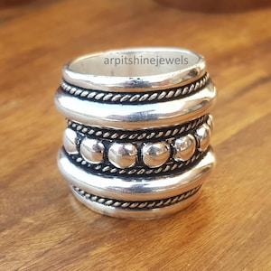 May include: A silver ring with a textured design. The ring features multiple bands with a rope-like texture and a row of small, round beads. The text "arpitshinejewels" is visible on the ring. The ring is on a wooden surface.