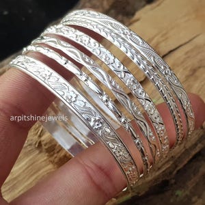 Op de afbeelding: Een stapel zilveren armbanden met ingewikkelde bloemen- en geometrische patronen. De armbanden zijn op een vinger gestapeld en tonen de gedetailleerde gravures. De merknaam "arpitshinejewels" is zichtbaar op de afbeelding.