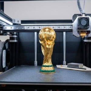 Puede incluir: Un trofeo dorado, similar al trofeo de la Copa Mundial de la FIFA, se encuentra sobre una plataforma de impresora 3D. La impresora es negra con rieles plateados y una bobina de filamento gris. Un pincel y un pequeño bloque gris están junto al trofeo.
