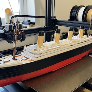Könnte beinhalten: Ein detailliertes Modell der RMS Titanic, mit schwarzem, rotem und weißem Rumpf und vier beigefarbenen Schornsteinen. Das Modell befindet sich auf einem 3D-Drucker, mit Filamentrollen in verschiedenen Farben darüber. Das Schiffsmodell ist etwa 76 cm lang.