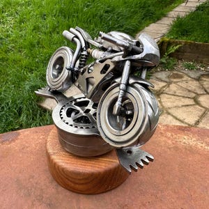 May include: A detailed metal sculpture of a motorcycle. The silver sculpture sits on a round, wooden base. The motorcycle features intricate details, including wheels, handlebars, and a seat. The sculpture is displayed outdoors.