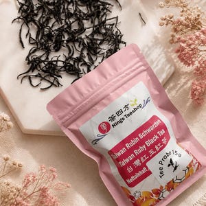 Könnte beinhalten: Eine rosa Teepackung mit dem Text "Taiwan Rubin Schwarztee" und "Taiwan Ruby Black Tea" neben einem Haufen loser schwarzer Teeblätter. Die Packung enthält auch den Text "Nings Teeshop" und "Probe 15g".