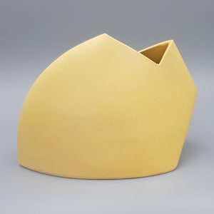 May include: A pale yellow ceramic vase with a smooth, matte finish and a distinct geometric silhouette. The object features a curved side that transitions into an angular, pointed top edge with a single triangular opening for floral arrangements.