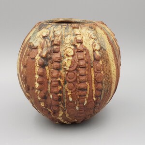 May include: A spherical ceramic vase featuring a textured surface with alternating vertical bands of reddish-brown clay and cream-colored glaze. The exterior is decorated with repeating embossed circular and rectangular patterns throughout the body.