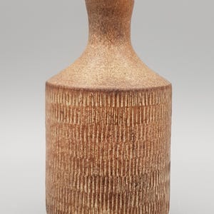 May include: A cylindrical ceramic vase with a narrow neck and a textured exterior featuring vertical incised lines. The surface has a mottled brown and tan finish with a matte appearance, showing a consistent pattern around the entire body.
