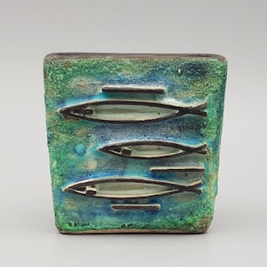 May include: A square ceramic vase with a textured surface featuring a gradient of blue and green glaze. The front displays three dark brown embossed fish silhouettes arranged horizontally, interspersed with small dark brown rectangular bars.