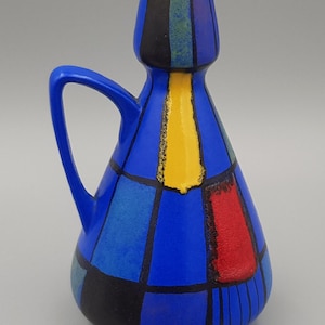 May include: A ceramic pitcher with a tapered neck and a single handle, featuring a vibrant blue base color decorated with a geometric pattern of black, yellow, and red rectangular blocks separated by thick black lines and a light blue interior rim.