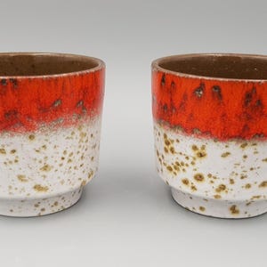 May include: Two ceramic cups featuring a vibrant orange glaze on the upper portion and a speckled white finish on the lower section. The interior of each cup is a solid brown colour, and the base of each vessel is slightly tapered and stepped.